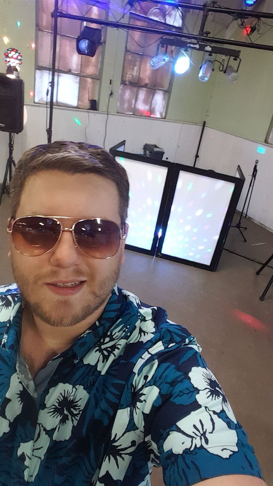 Adam Plett in sunglasses and a blue patterned shirt with his DJ setup behind him